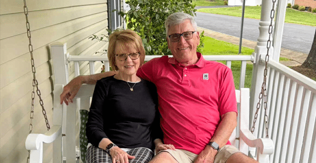 Joan and Monty Montgomery, an older adult couple, sit side by side on a white porch swing. Joan is wearing a black top and plaid pants, while Monty is in a pink polo and khaki shorts. They are smiling and relaxed on the covered porch of their Sunnyside home, with white railings and greenery in the background.