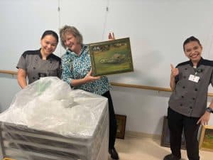 Sunnyside staff members laughing and working together to hang resident artwork in Artist Alley, a creative gallery space inside Sunnyside Retirement Community.