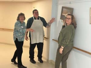 Sunnyside staff members laughing and working together to hang resident artwork in Artist Alley, a creative gallery space inside Sunnyside Retirement Community.