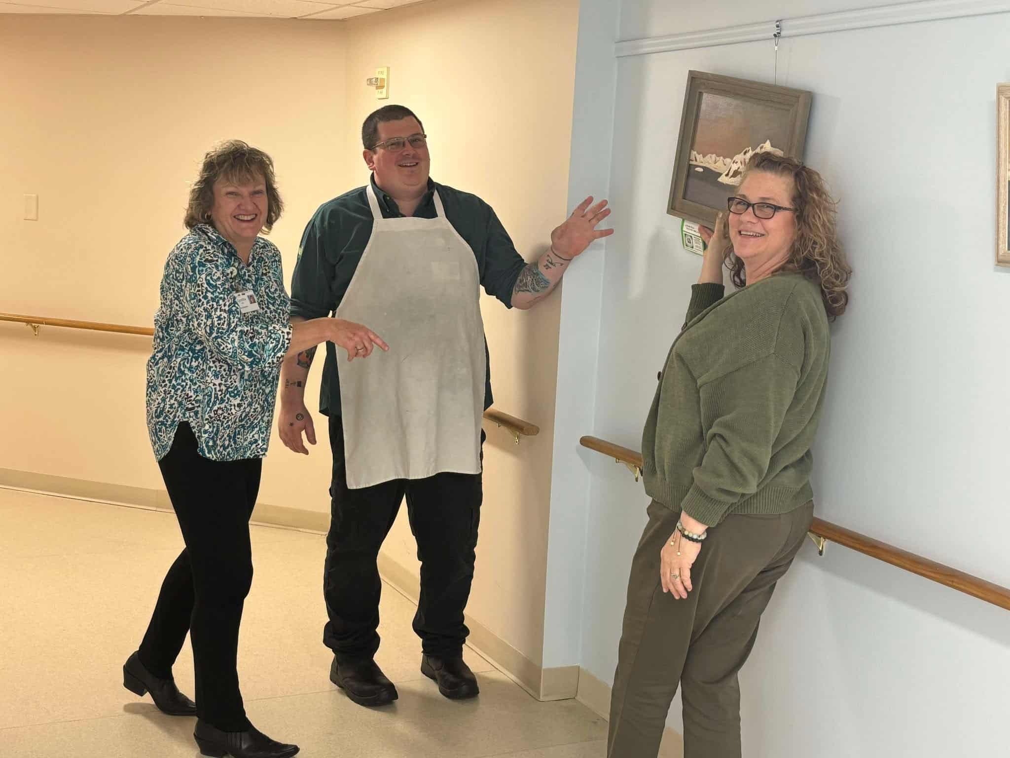 Sunnyside staff members laughing and working together to hang resident artwork in Artist Alley, a creative gallery space inside Sunnyside Retirement Community.