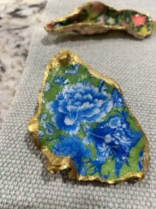 A hand-painted oyster shell decorated with a bold blue floral design on a green background, outlined in metallic gold. The shell rests on a light gray woven fabric with another painted shell blurred in the background.