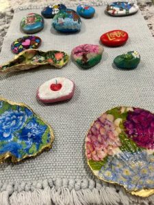 A collection of brightly painted rocks and oyster shells displayed on a textured gray woven cloth. The rocks are decorated with colorful designs including hearts, animals, flowers, and words like “LOVE.” Two oyster shells near the front are painted with vivid floral patterns and trimmed with gold edges.