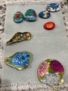 A hand-painted scallop shell featuring a vibrant floral collage with pink, purple, and blue blossoms, finished with a glossy seal and a gold-painted rim. The shell rests on a light gray woven cloth with fringe, atop a speckled countertop.