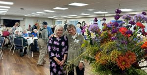 Residents Judith Anderson and Barb Mancini stand together smiling beside a vibrant flower arrangement at the Sunnyside Art Gala.