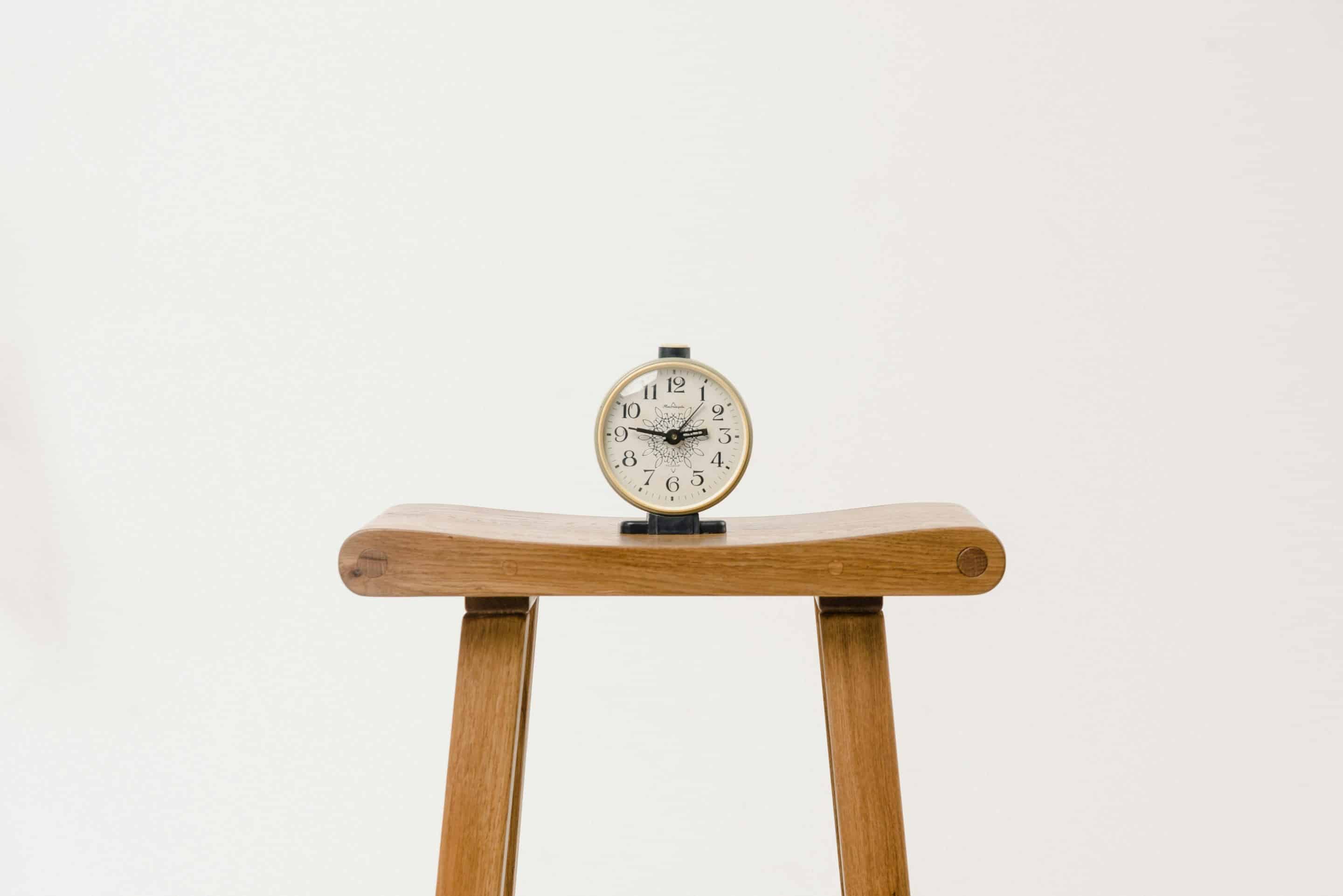 time "Vintage-style alarm clock with black numbers and ornate clock hands, placed on a minimalist wooden stool against a plain white background."