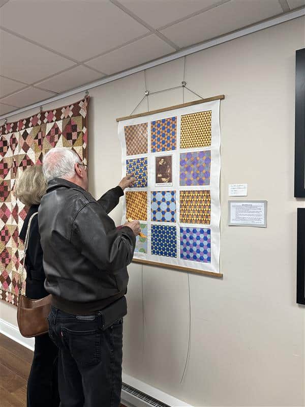 A couple studies a quilted wall hanging featuring geometric patterns and bright colors.