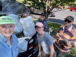 Four residents smile while standing beside a vehicle loaded with large bags of yard debris. They're dressed for outdoor work, wearing hats, gloves, and sunglasses after a successful cleanup effort on a sunny day.