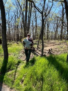 A man stands in a sunlit forest clearing, holding a bag of gathered yard debris. Bright green grass and tall trees surround him, capturing the beauty of a spring morning cleanup.