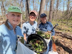 Three women smiling and standing in a wooded area on a sunny day, participating in a cleanup effort. They are holding buckets and bags filled with leaves and plants, wearing gloves, hats, and casual outdoor clothing. Trees with early spring foliage surround them.