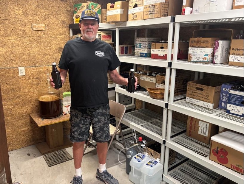 Ed Adams A man wearing a black T-shirt, camouflage shorts, and a "Beer" cap stands in a storage room holding two dark beer bottles. Behind him are shelves lined with labeled boxes and brewing supplies, and a large glass jug filled with liquid sits on a small table.