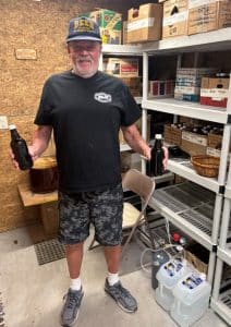 A man wearing a "Beer" cap, black T-shirt, and camouflage shorts stands in a storage room holding two dark beer bottles. Behind him are shelves filled with labeled boxes and brewing supplies, and a large glass container of fermenting beer sits on a small wooden table.